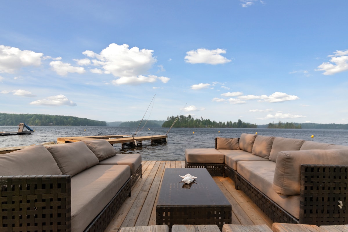 A spacious outdoor seating area features comfortable sectional sofas arranged around a low coffee table. The setting overlooks a tranquil lake surrounded by greenery, with a wooden dock visible in the distance under a bright sky dotted with fluffy clouds.