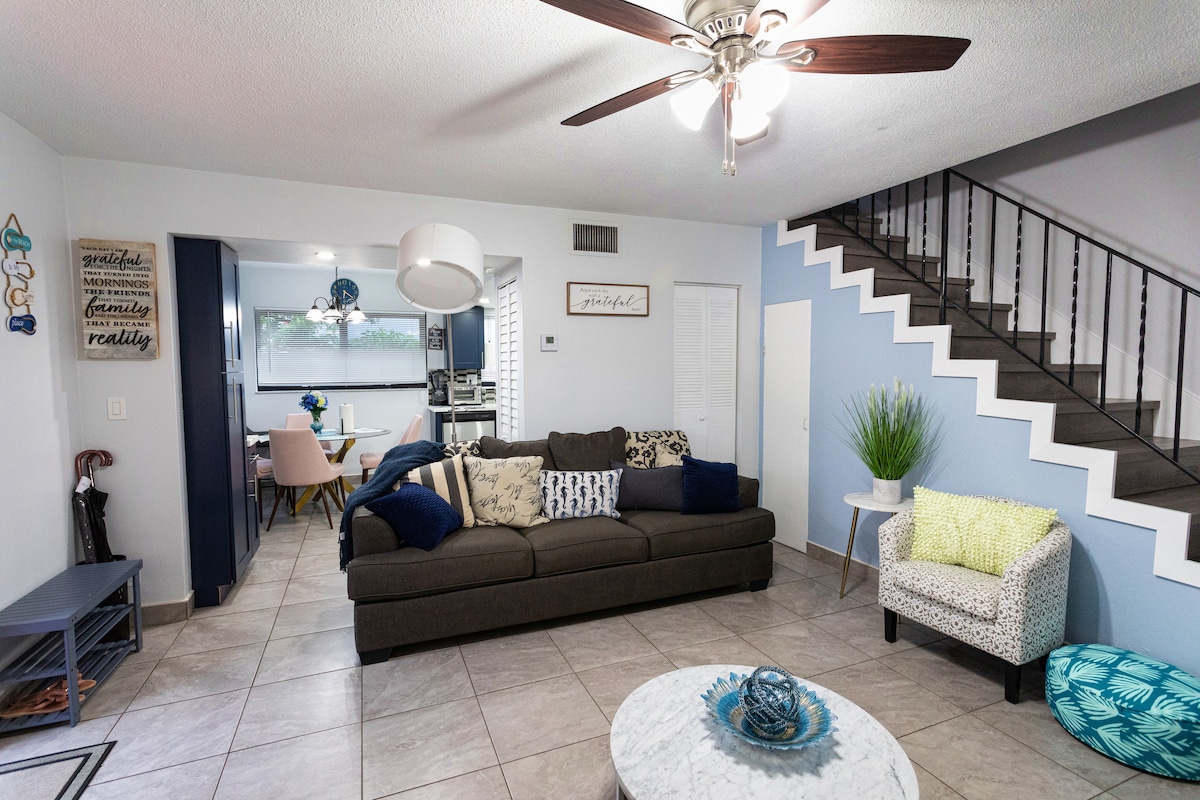 A spacious living room features a dark gray sofa with decorative pillows, a white marble coffee table, and a ceiling fan. A light fixture hangs above, while a staircase leads to the upper level. The open layout connects to a dining area in the background.