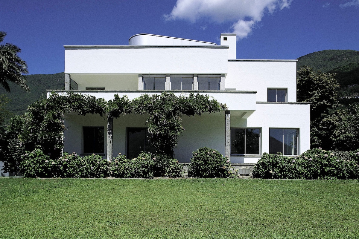 The modern white facade of the house is complemented by lush greenery and vines enveloping the terrace. Large windows reflect the surrounding nature, while the expansive lawn provides an inviting space for relaxation and outdoor activities.