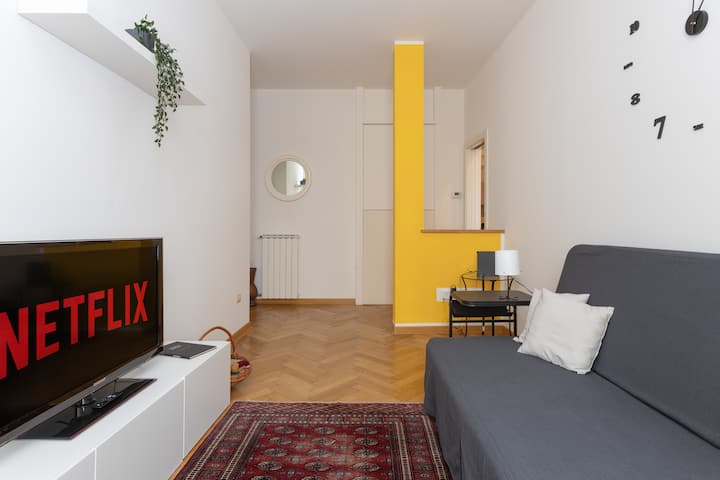 Zonë ndenjeje me divan-krevat, dollap të bardhë me Smart TV, tapet, një kolonë ndarëse të verdhë dhe një orë muri.