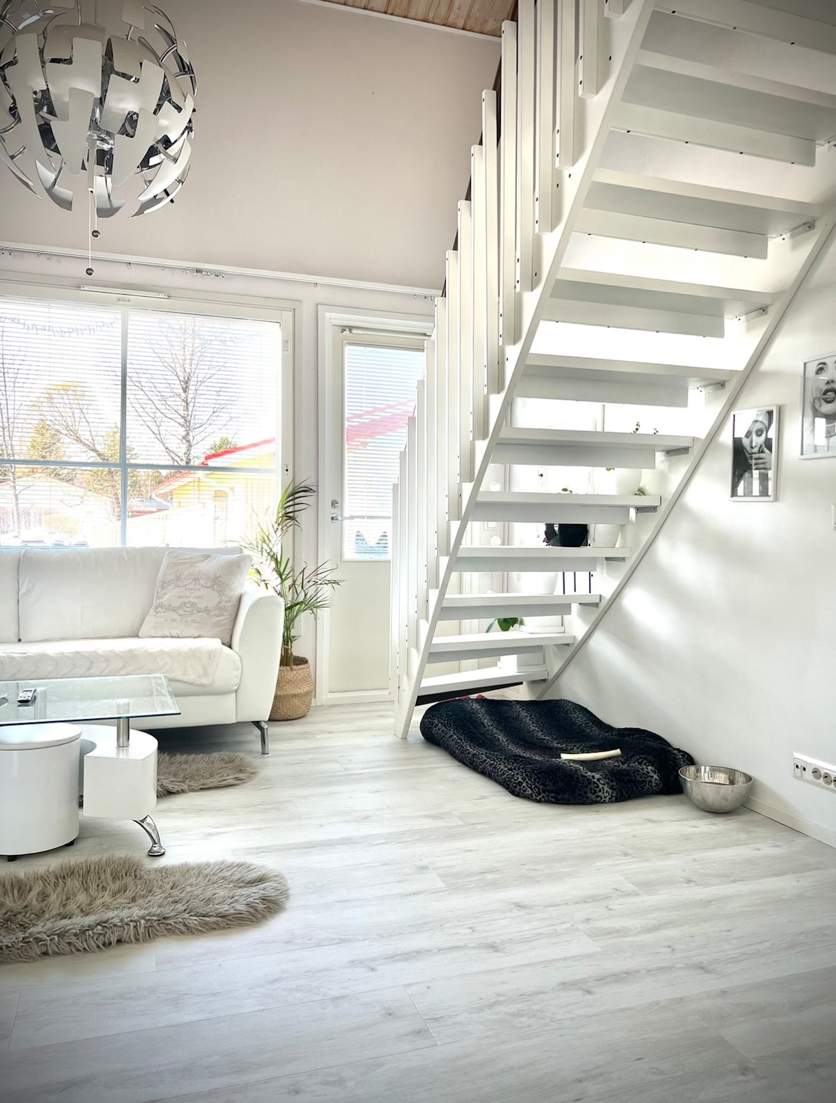 A bright living area features a white sofa paired with a glass coffee table. Natural light flows through large windows, illuminating a minimalist décor. A modern staircase leads to an upper level, while a cozy pet bed is positioned nearby, enhancing the inviting space.