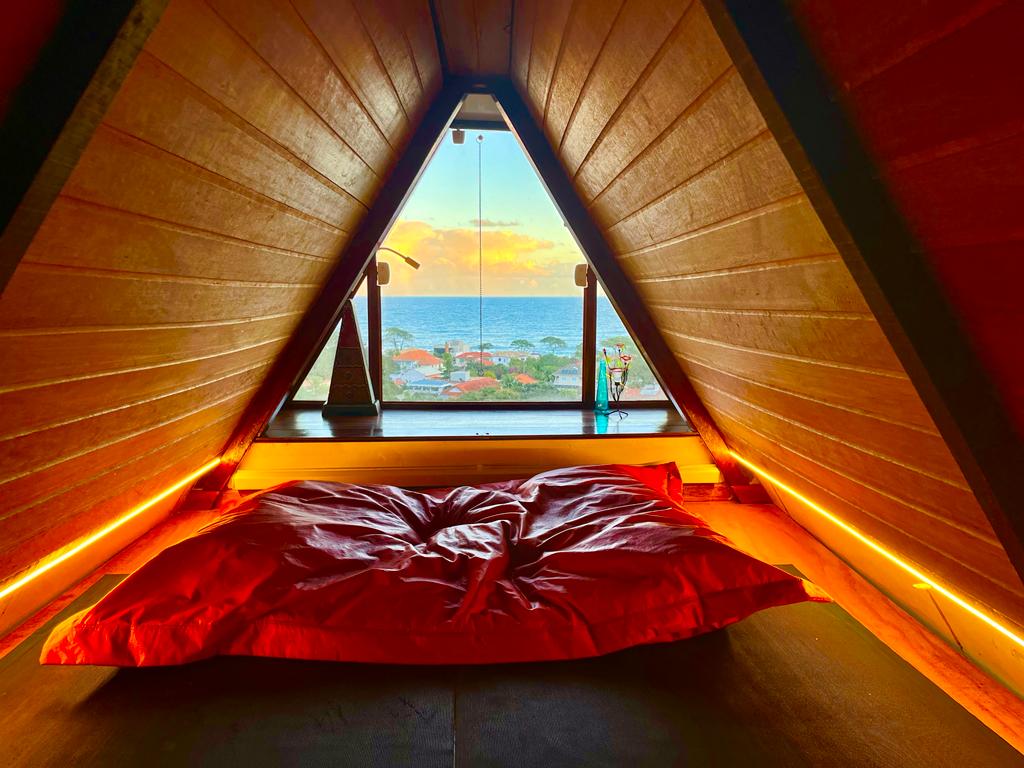 An angular loft space features a large, bright red cushion positioned near a window, offering views of the ocean and sky. Warm lighting illuminates the wooden walls, creating a cozy and inviting atmosphere. The scene reflects a tranquil setting ideal for relaxation.