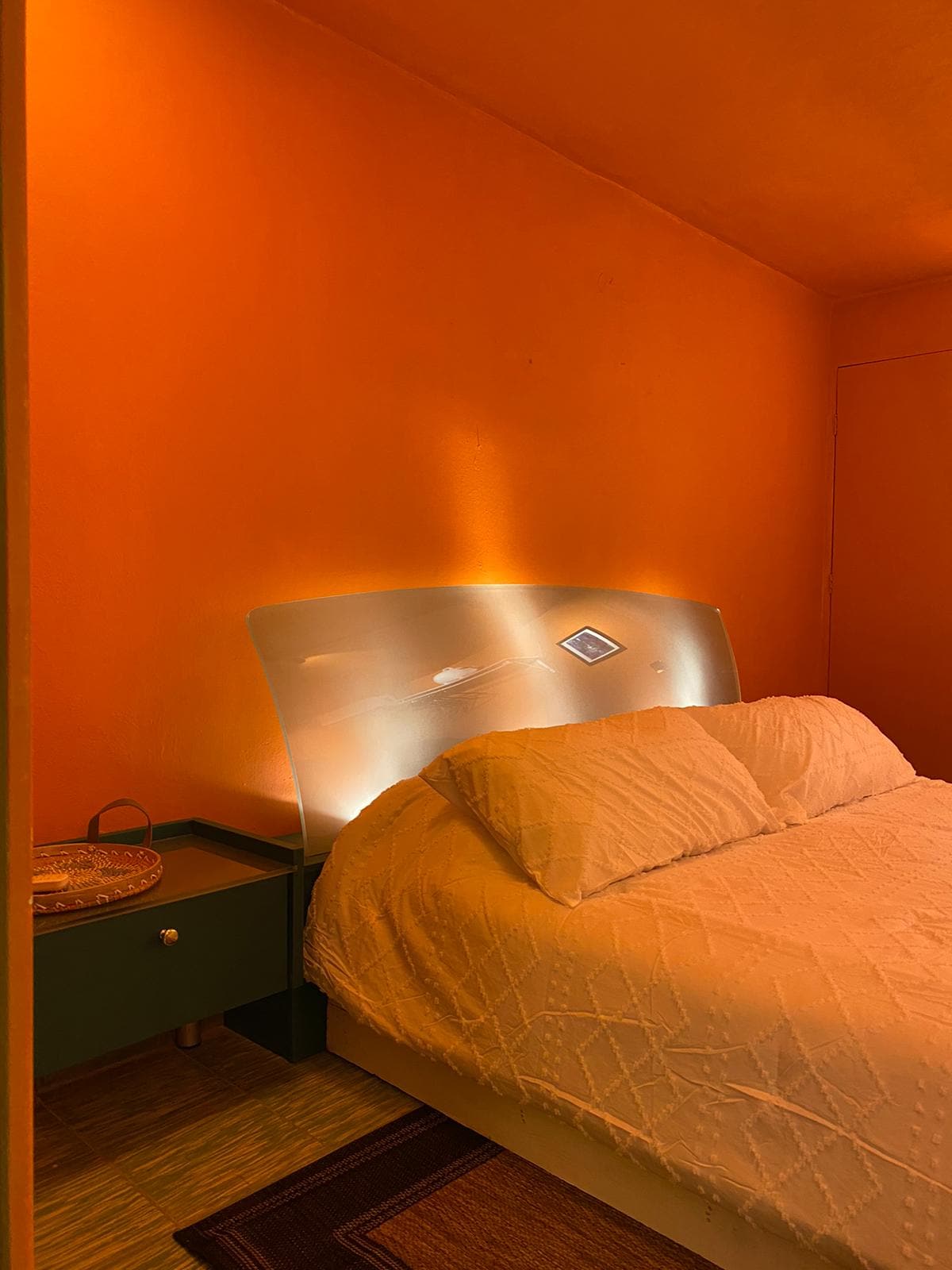 A vibrant orange room features a spacious bed with a textured white cover, complemented by a sleek headboard that emits soft lighting. A small green nightstand with a decorative tray is seen beside the bed, enhancing the room's bright and modern aesthetic.