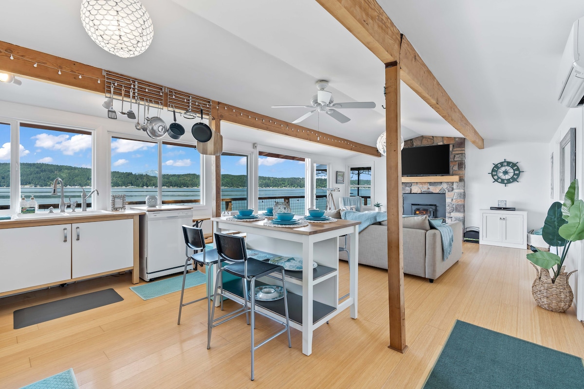 The open-concept living area features a modern kitchen with hanging cookware and a dining table set for meals. Large windows provide scenic views of the surrounding landscapes, while minimalist furnishings create a clean and welcoming atmosphere. A stone fireplace adds warmth to the space.
