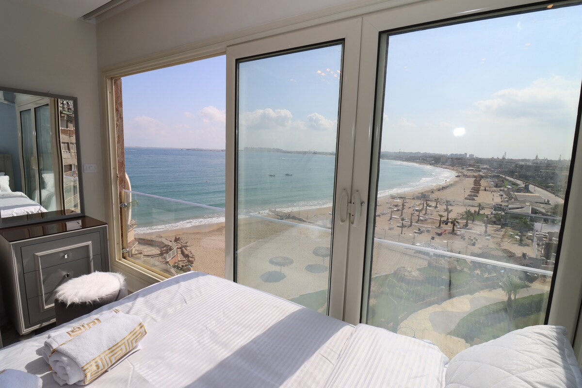 A spacious sleeping area features a large bed positioned near floor-to-ceiling windows. Stunning views of the Mediterranean Sea and sandy beach are visible, with gentle waves lapping at the shore. Soft natural light brightens the room, enhancing the tranquil ambiance.