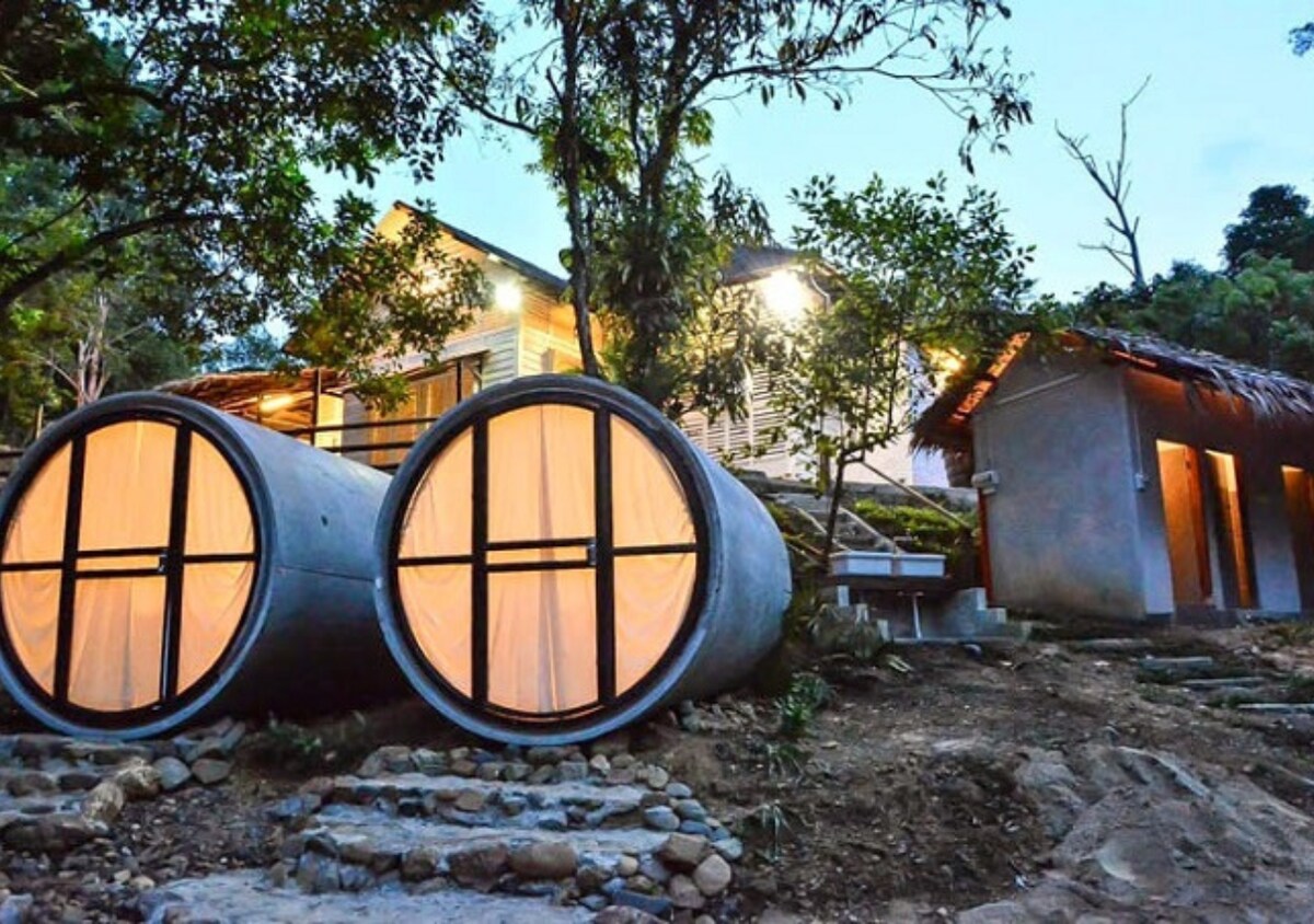 Two unique cylindrical accommodations are positioned amidst a natural landscape, with warm light glowing from within. A rustic building with a thatched roof is visible nearby, surrounded by greenery and stone pathways that lead through the tranquil setting.