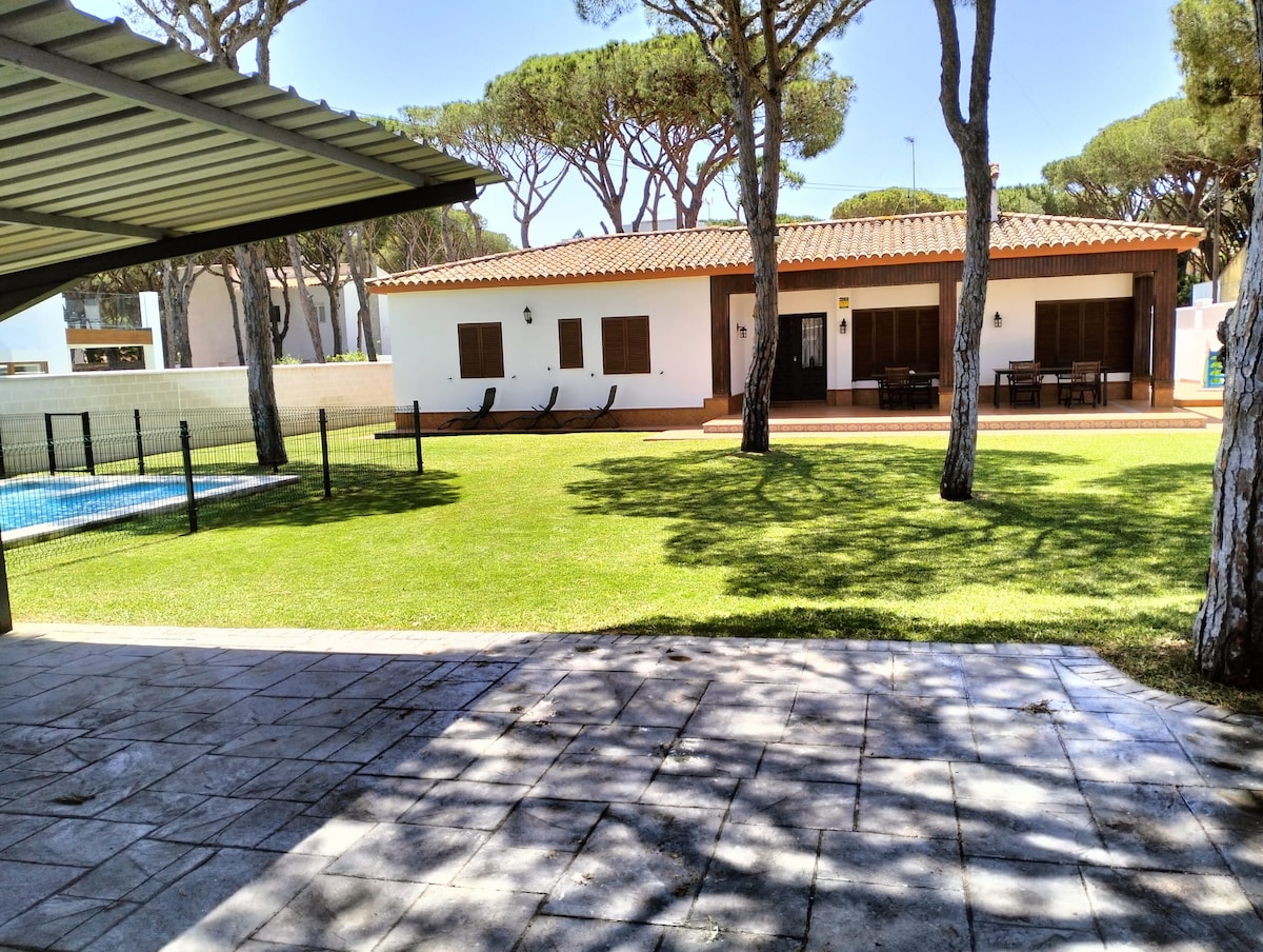 A spacious outdoor area features a well-maintained lawn, with a view of a single-story building that includes large windows and a terracotta roof. A shaded patio area is visible beside a private pool, with lounge chairs positioned nearby under tall trees.