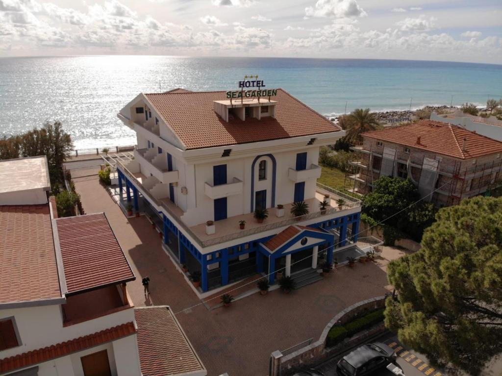 The Sea Garden Hotel is depicted from an aerial view, showcasing its modern architecture and welcoming facade. The building is surrounded by palm trees and outdoor seating, with a glimpse of the beach and ocean in the background under a partly cloudy sky.