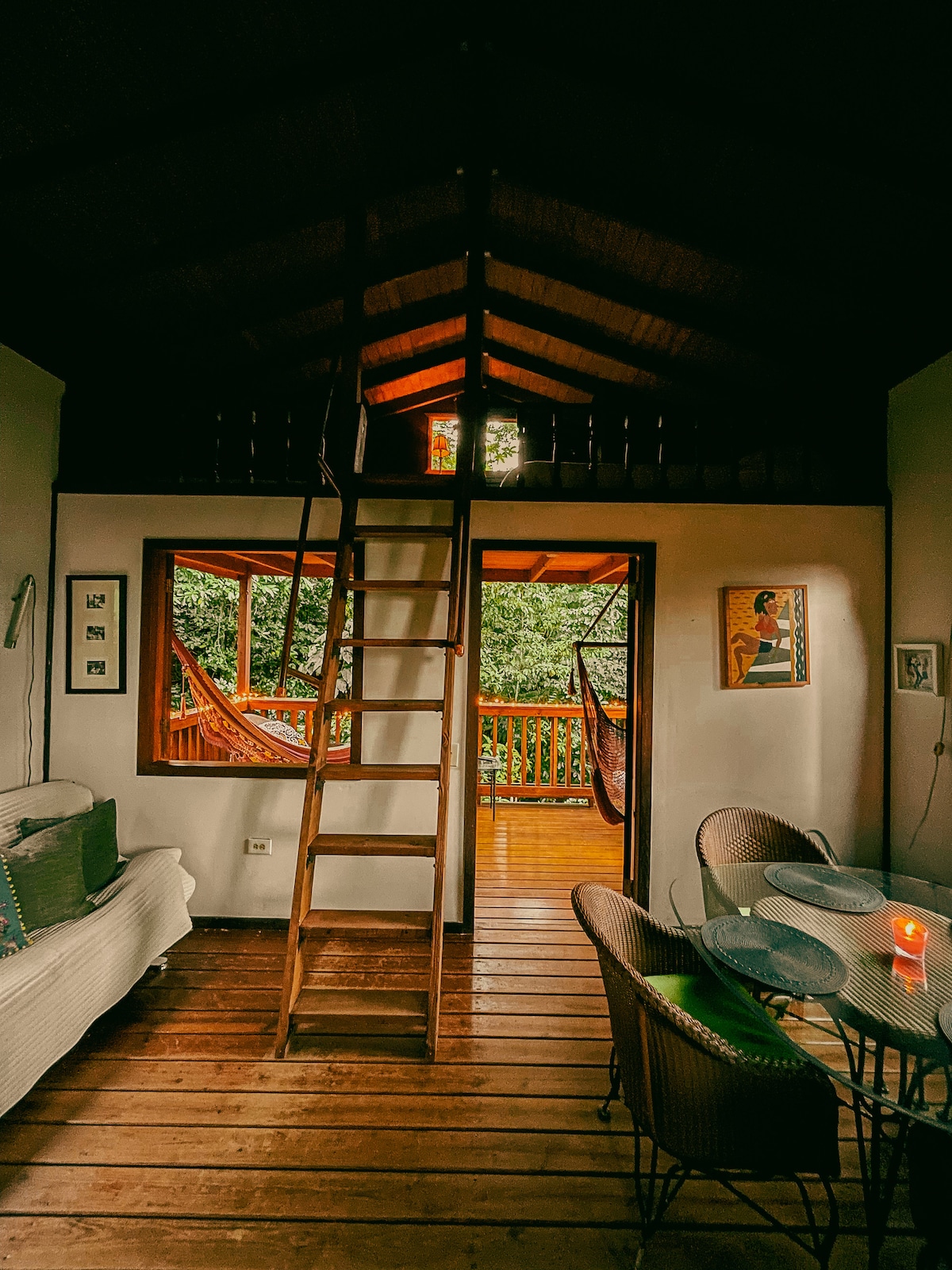 An interior view of a rustic wooden loft features a staircase leading to an upper level. Natural light filters through windows, highlighting a seating area with a white sofa and a dining table with wicker chairs. A hammock is positioned on the balcony outside, surrounded by lush greenery.
