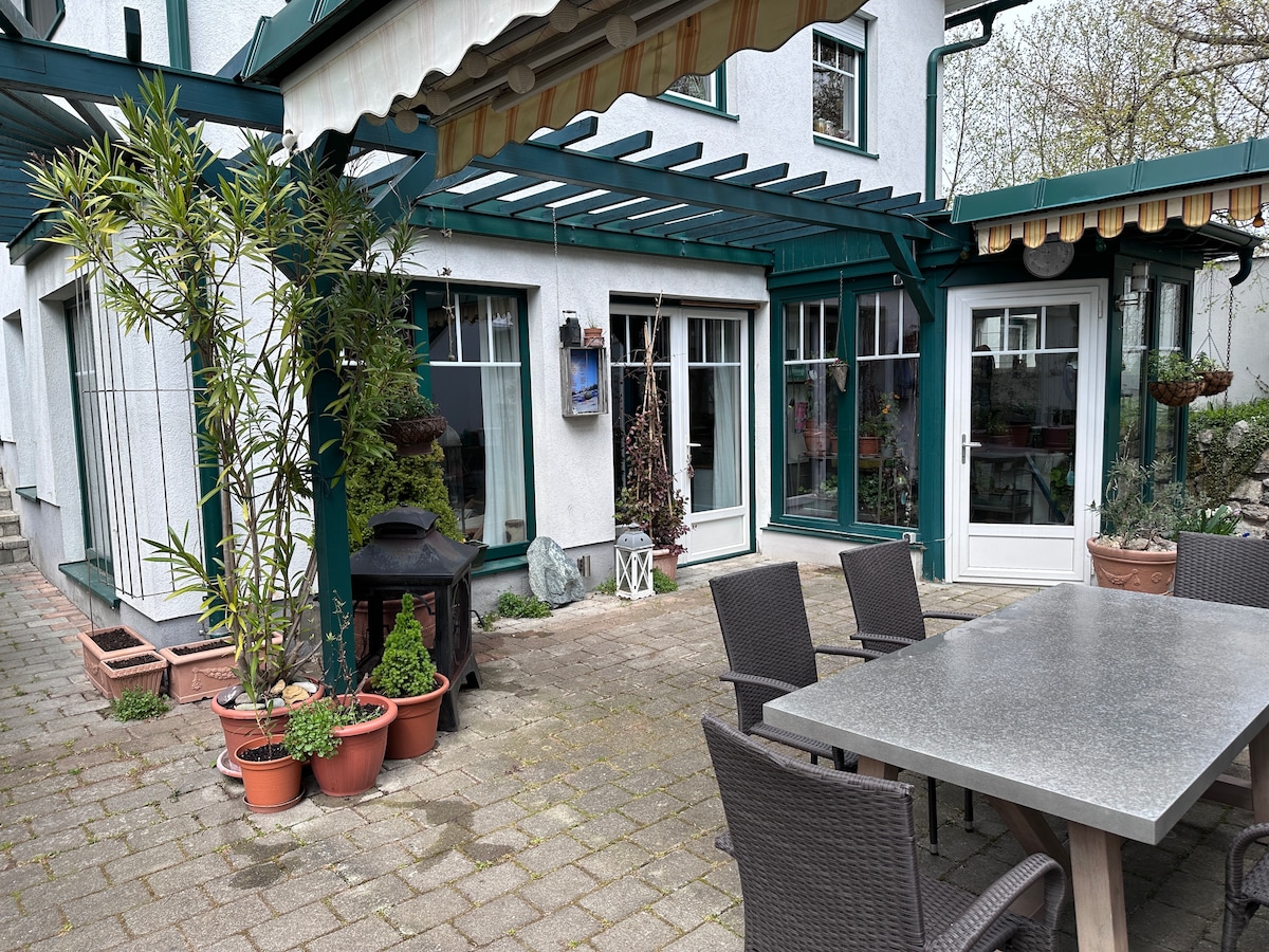 An outdoor patio area is presented, featuring a large stone table surrounded by multiple chairs. Potted plants add greenery to the space, while a grill is positioned nearby. Light filters through the awning, enhancing the openness of the terrace's design.