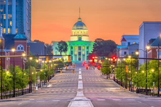 A view of a wide street leading towards a large, illuminated building with a dome, surrounded by trees. The sky is painted in soft hues of orange and blue as dusk approaches. Streetlights line the road, contributing to the evening ambiance.