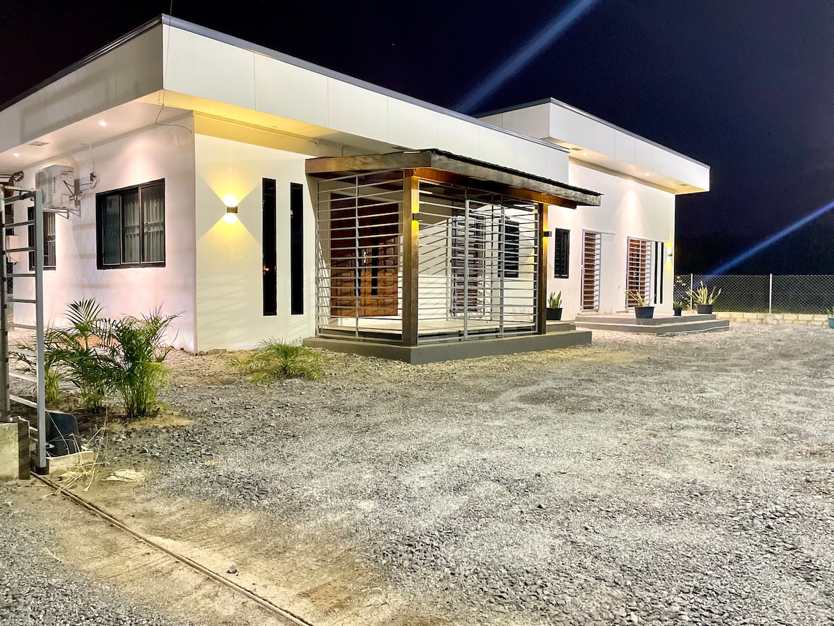 The exterior of a modern single-story building is shown at night, featuring a minimalist façade with large windows. A covered porch area is visible, surrounded by a gravel driveway and planters with foliage. Soft lighting enhances the clean lines of the architecture.