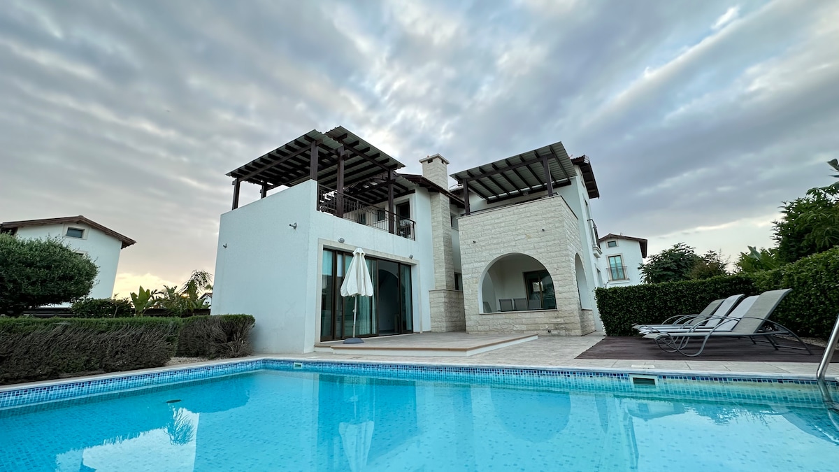 A modern villa is showcased with a spacious patio overlooking a private pool. Comfortable sun loungers line the pool deck, while an outdoor dining area is shaded by a wooden pergola. A lush garden and the Mediterranean backdrop complete the serene scene.