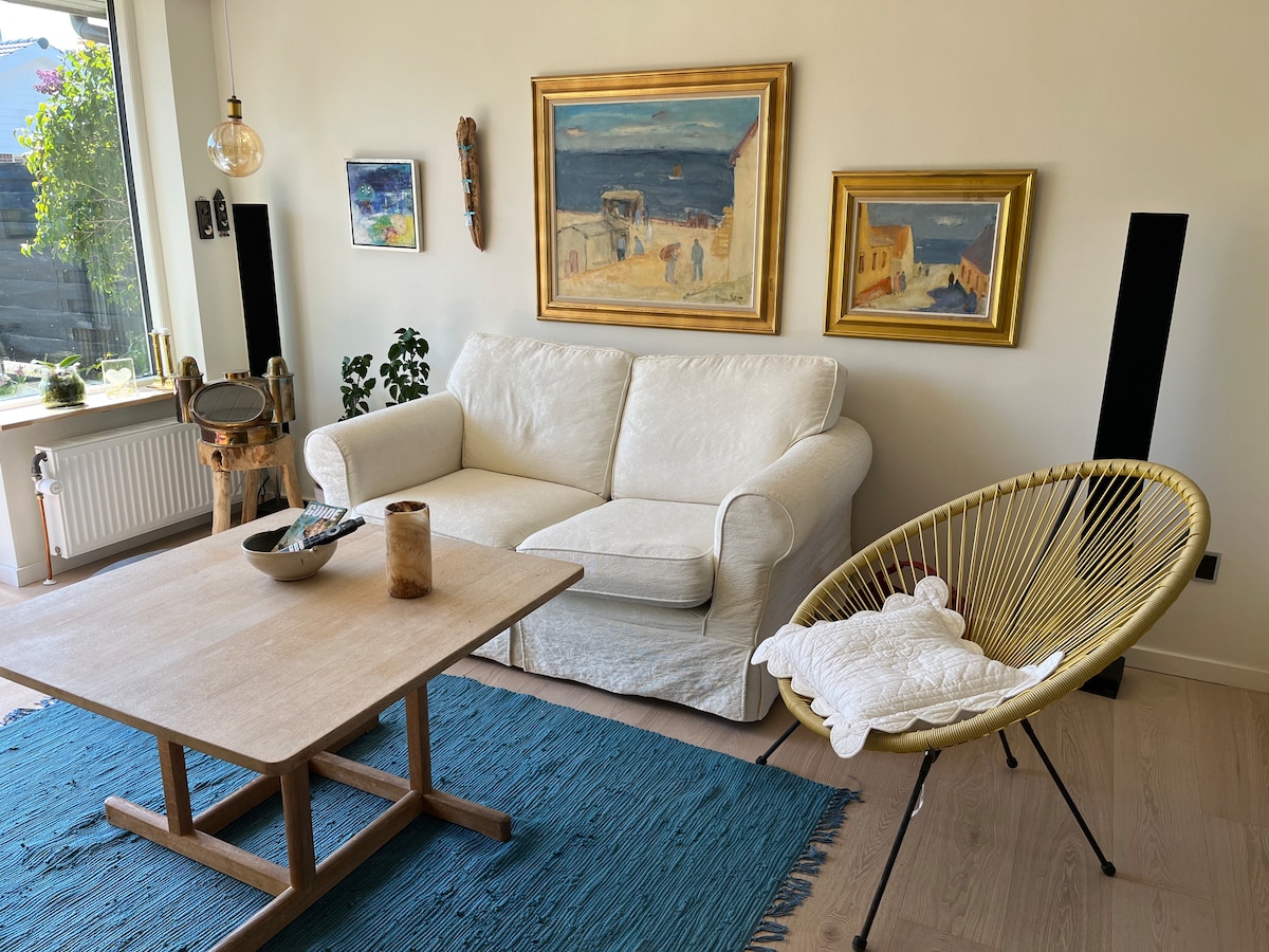 A bright living area is highlighted by a light-colored sofa and a round, woven chair. The wooden coffee table sits on a textured blue rug, surrounded by potted plants. Two framed paintings adorn the walls, enhancing the peaceful ambiance.