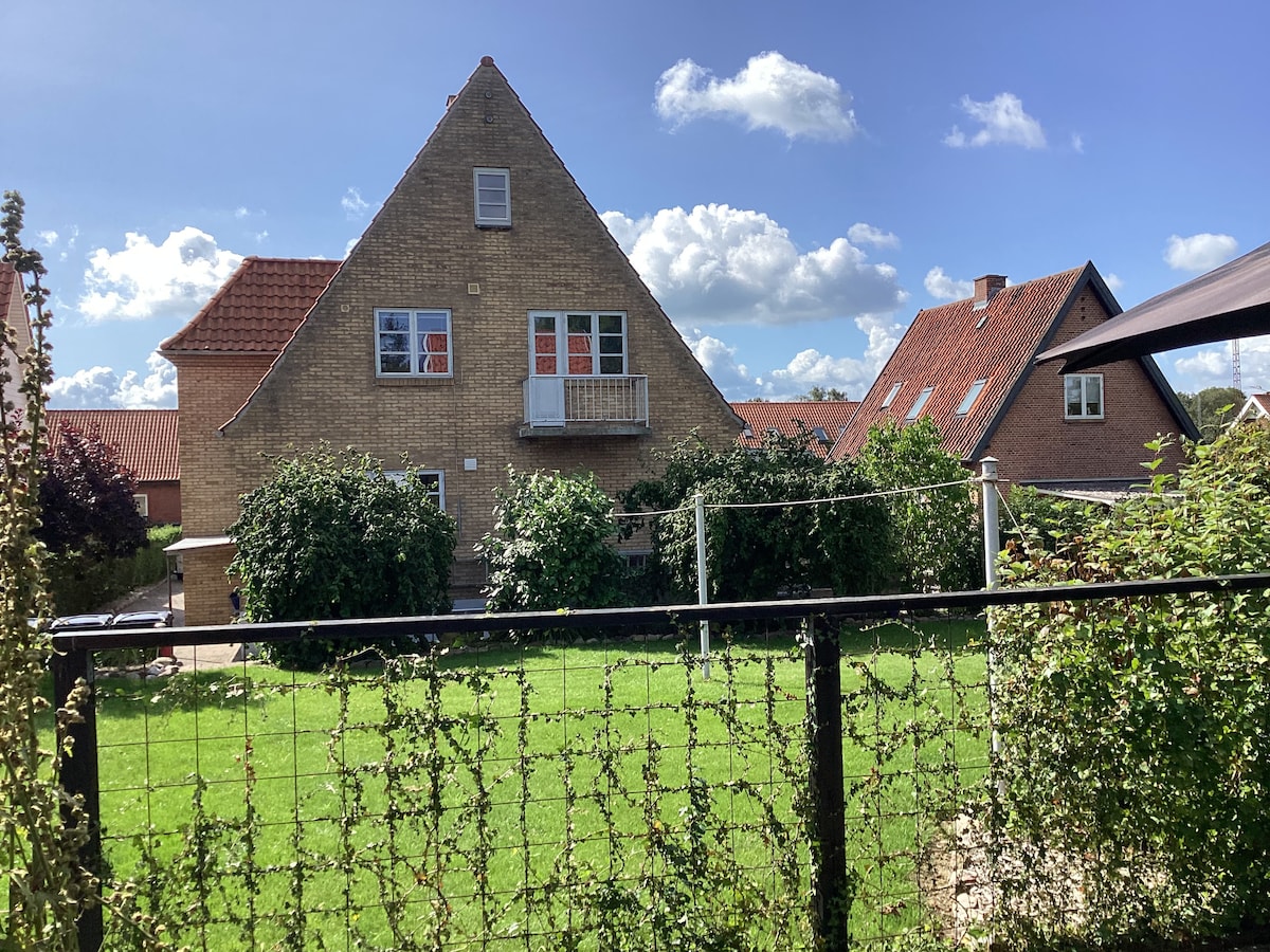 A two-story building is surrounded by lush greenery, featuring a sloped roof and multiple windows. A balcony is visible on the upper floor, while the ground is lined with well-maintained grass. The sky is clear with scattered clouds, contributing to a serene outdoor setting.