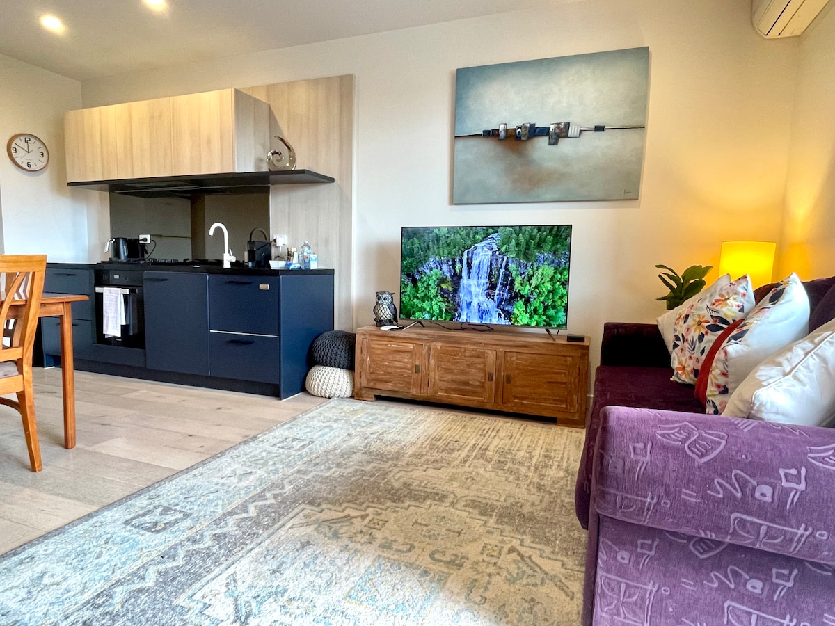 An open-plan living area is visible, featuring a modern kitchen with dark cabinetry and a large flat-screen TV displaying a nature scene. A comfortable sofa, decorated with patterned cushions, complements a wooden dining table with chairs. Soft lighting creates a welcoming atmosphere.