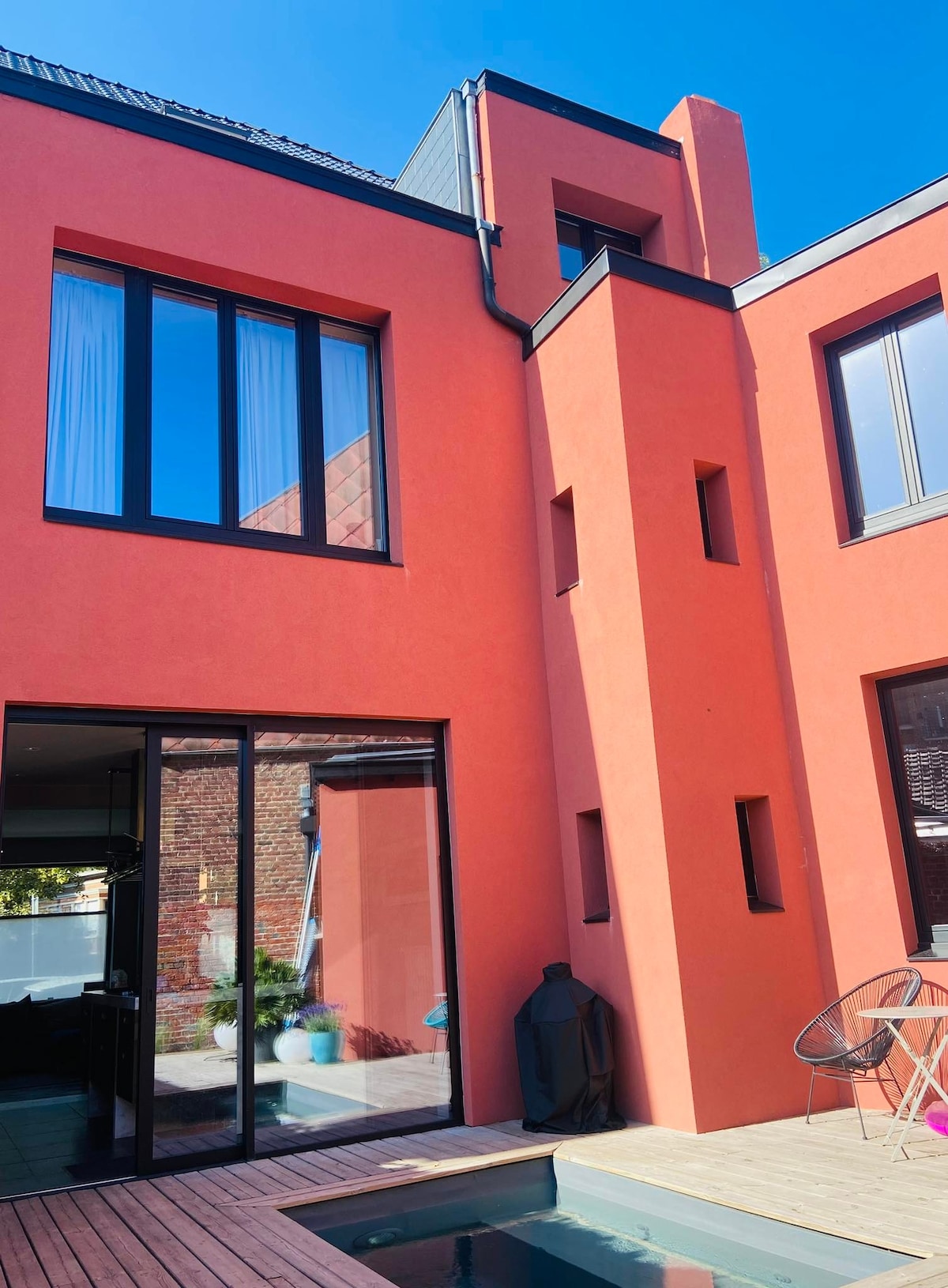 The exterior of a renovated house is showcased in a vibrant orange hue. Large glass windows reflect the sunlight, offering views into the interior. A sleek modern pool is visible in the foreground, complementing the contemporary architecture.