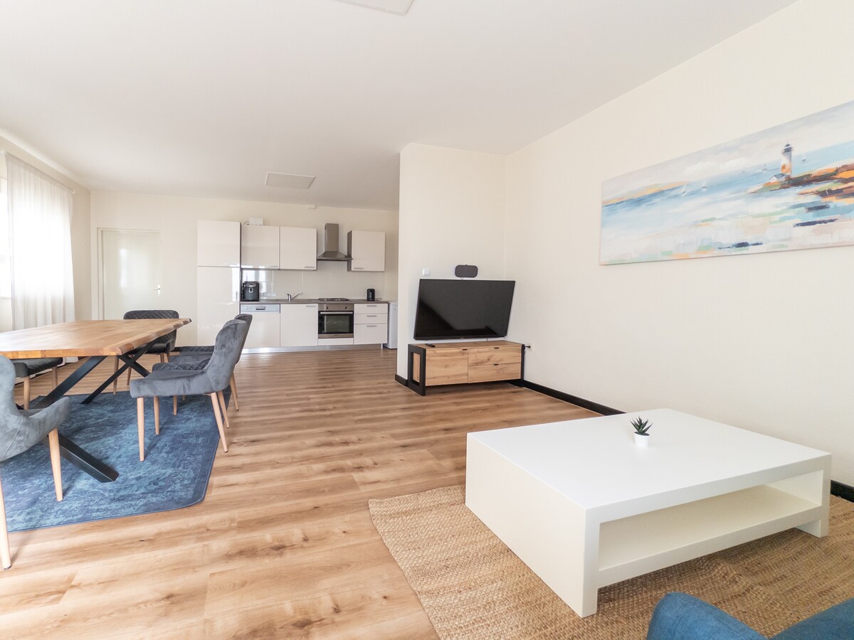 An open living area features a large dining table surrounded by gray chairs. A modern kitchen is visible in the background, equipped with stainless steel appliances. A white coffee table sits on a natural fiber rug, and light streams through large windows, enhancing the bright atmosphere.