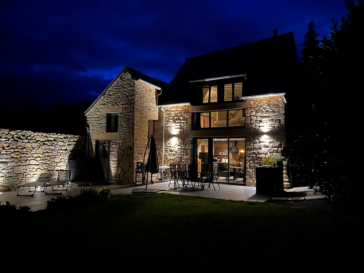 The exterior of the renovated stone house is illuminated at night, showcasing its charming architecture. Soft lights highlight the structure and seating area, creating a serene atmosphere in the garden. A patio with chairs and tables invites relaxation under the evening sky.