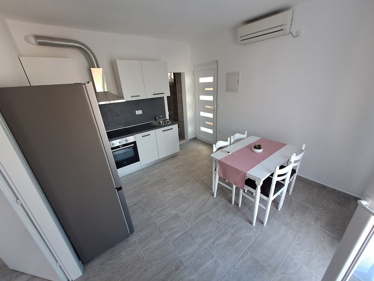 A modern kitchen area is showcased, featuring white cabinetry and stainless-steel appliances. A dining table with six white chairs occupies the space, complemented by a soft pink table runner. A door with frosted glass panels allows natural light to fill the room.