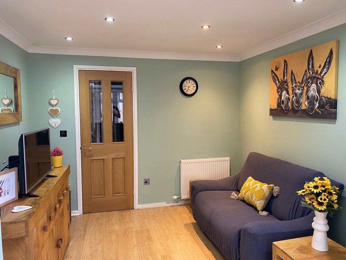 A cozy living area features light green walls and wood flooring. A comfortable sofa is positioned against the wall, accompanied by a decorative cushion. A wooden sideboard and clock add functionality, while artwork of donkeys enhances the space's character.