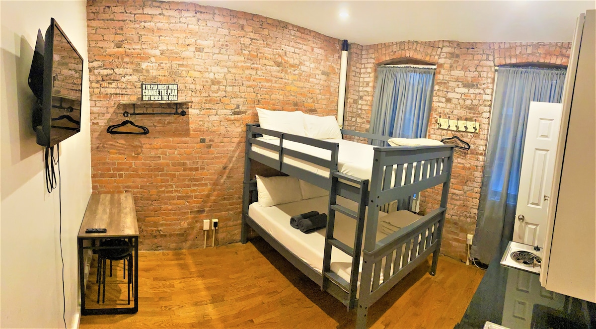 A brick-walled room features a sturdy bunk bed with a spacious lower and upper sleeping area. A small wooden table with stools is positioned nearby, and a flat-screen TV is mounted on the wall. Natural light filters through sheer curtains at the window.