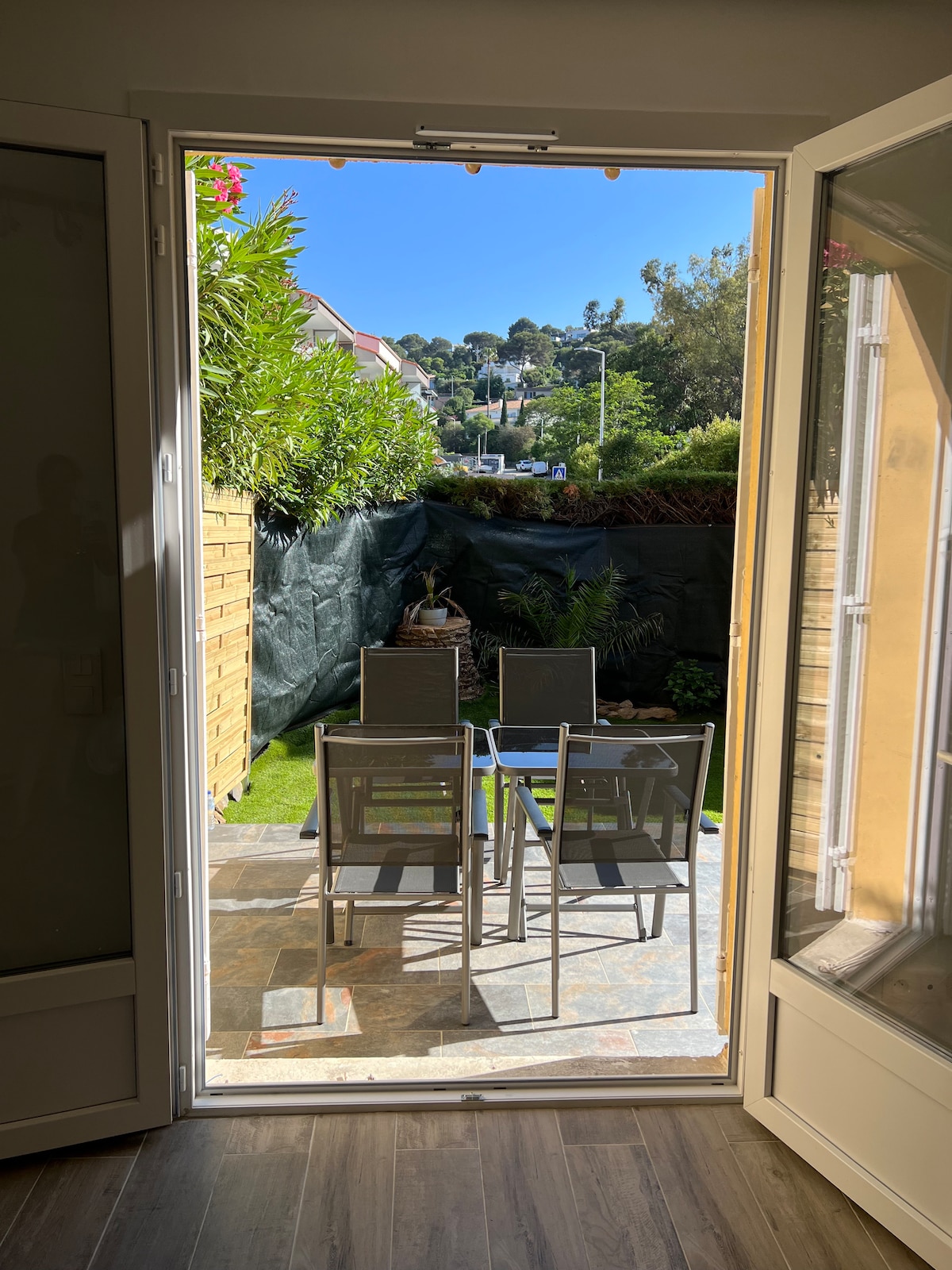 A bright outdoor terrace is visible, featuring a table and four chairs set on stone flooring. Lush greenery surrounds the space, while a sunny view of distant hills is observed through open glass doors.