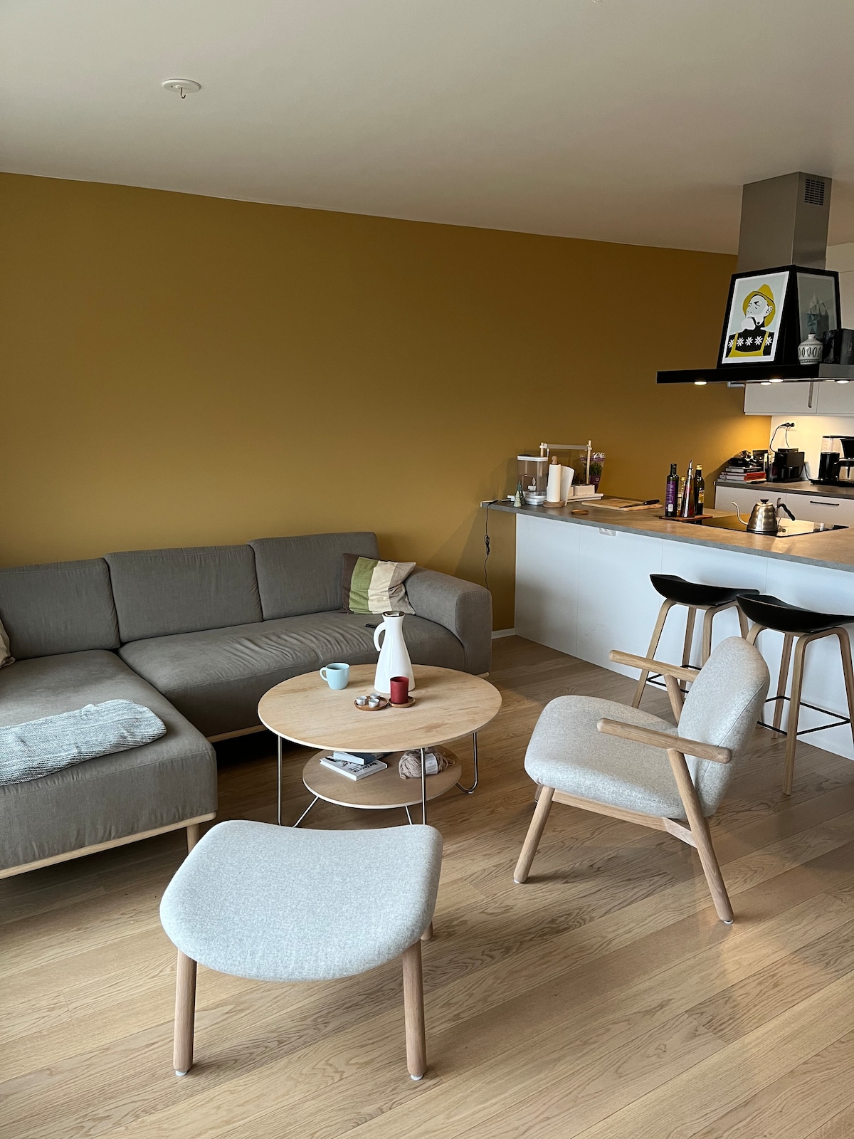 A modern living area is depicted with a cozy gray sofa, complemented by light wooden flooring. A round coffee table is positioned center, holding various items. In the background, a sleek kitchen can be seen featuring a minimalist design and modern appliances.