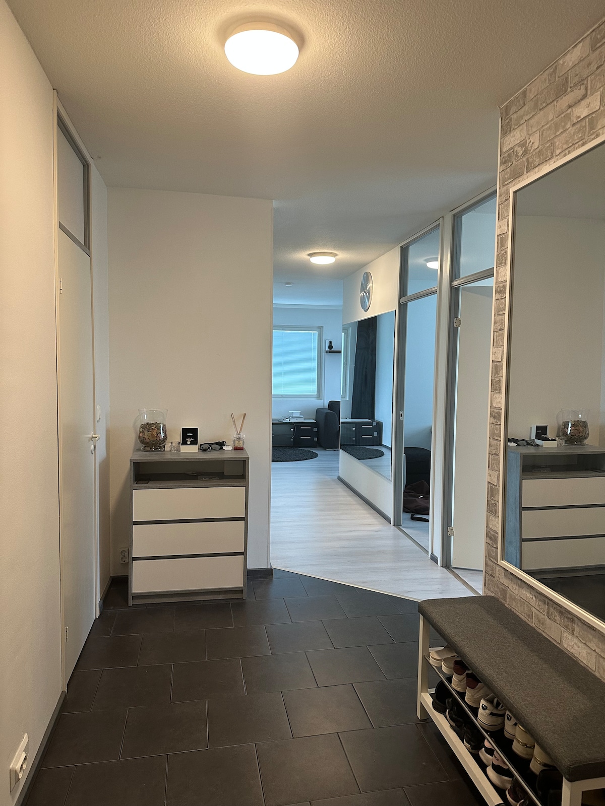 The entrance area features a sleek tile floor, complemented by a shoe storage bench and a mirrored cabinet. A spacious office room with glass doors is visible ahead, showcasing a desk and chair arrangement. Natural light filters in from a nearby window.