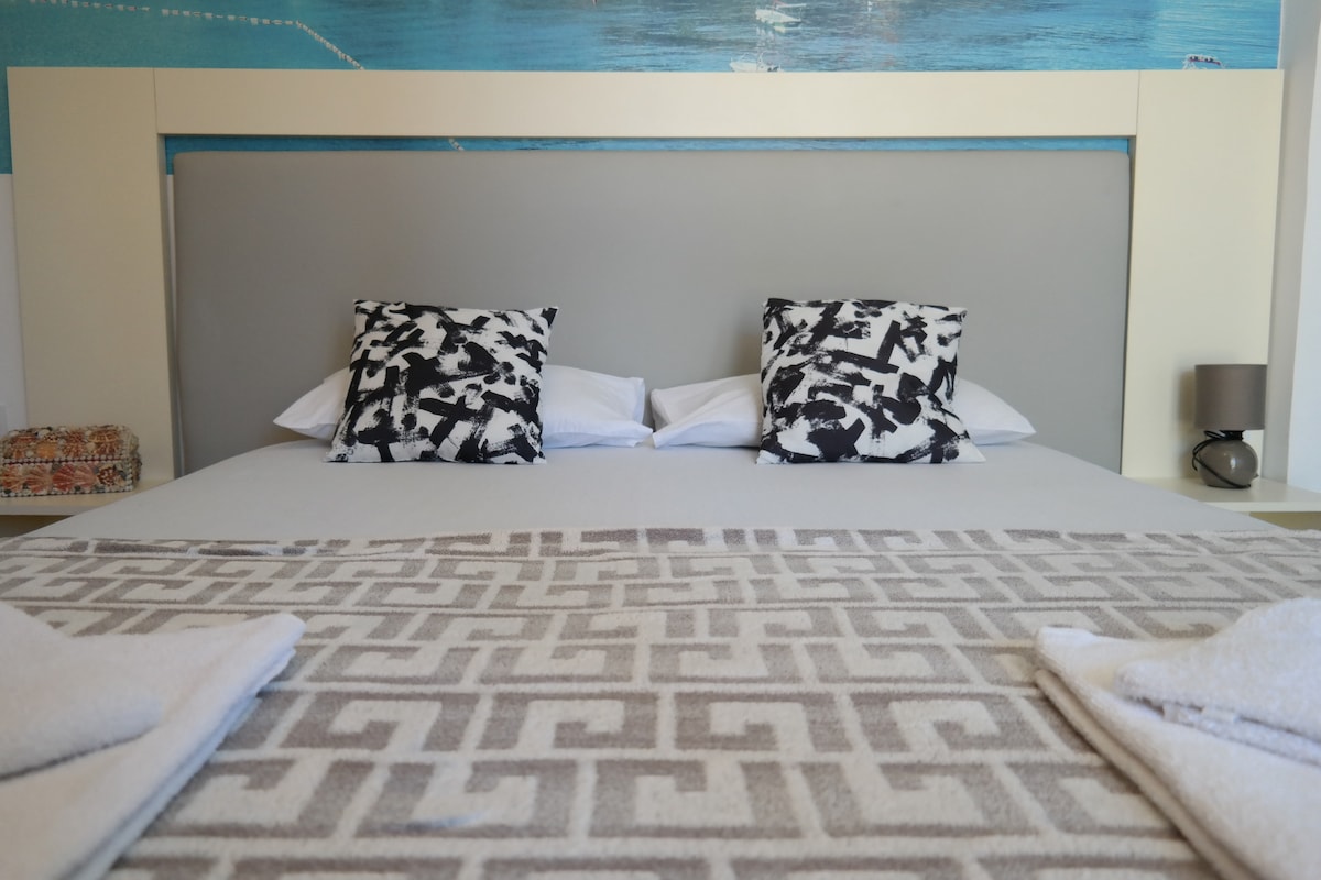 A spacious bed is centered in the room, adorned with decorative black and white pillows. A soft, patterned blanket is draped across the bed, and towels are neatly positioned at the foot. A bedside lamp is visible, contributing to the overall cozy ambiance.