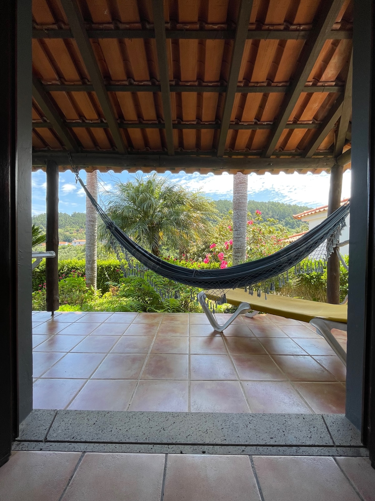 A private terrace offers a view of lush greenery and distant hills. A comfortable hammock is suspended between wooden posts, with a sun lounger positioned nearby. The tiled floor and wooden ceiling create a relaxed outdoor space, perfect for enjoying the surrounding nature.