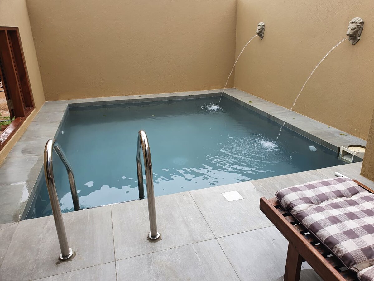 A private plunge pool is featured, surrounded by smooth stone tiles with light splashes created by fountains. Two elegant wall-mounted sculptures enhance the serene environment. A wooden lounge chair with a patterned cushion is positioned nearby, inviting relaxation beside the refreshing water.