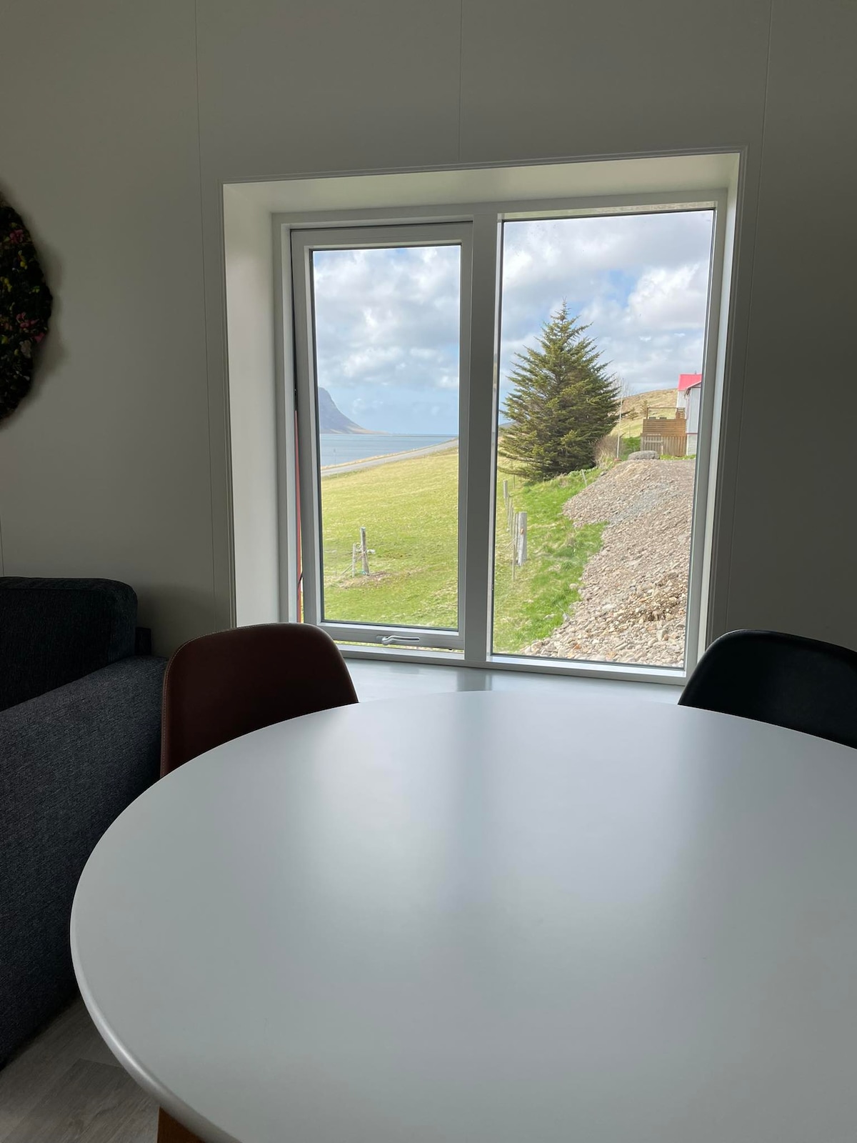 A round white dining table is positioned near a large window, offering views of the grassy landscape and distant mountains. Natural light fills the space, highlighting the serene setting just outside the window.