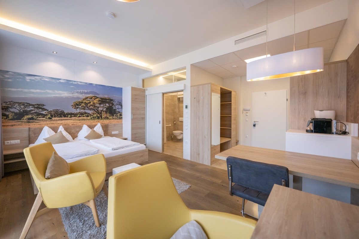 A bright and airy studio features a spacious bed framed by light wooden elements. A vibrant mural of a savanna landscape enhances the ambiance. A small kitchenette, dining table, and yellow armchairs provide functional seating, while modern bathroom facilities are accessible through a sliding door.