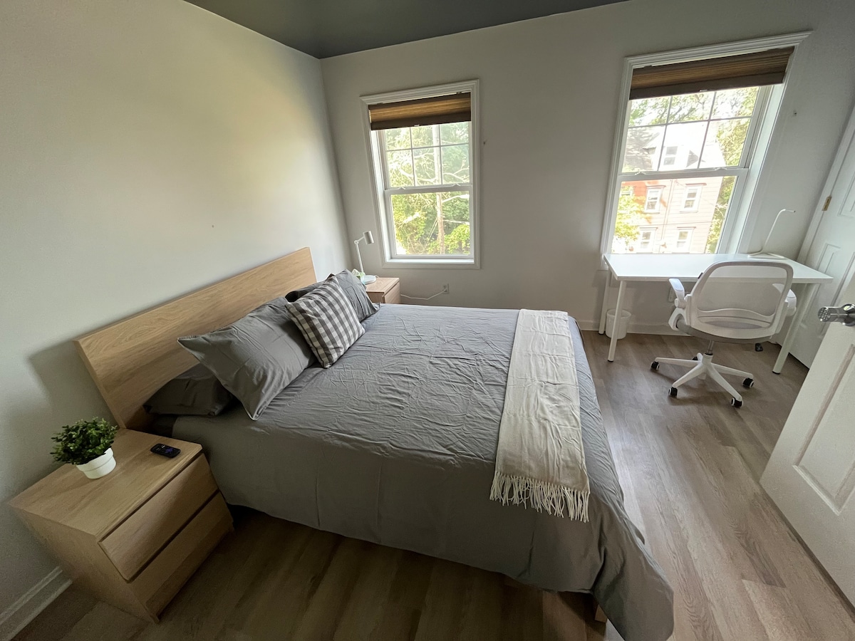 A well-appointed bedroom features a spacious bed with gray linens and decorative pillows. Natural light illuminates the room through two windows with wooden shades. A desk and chair provide a dedicated workspace, while light wood nightstands complement the overall decor.
