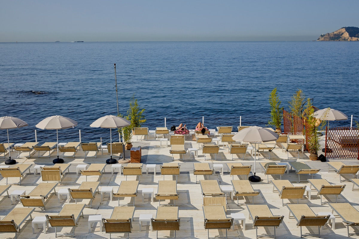 A spacious sun terrace overlooking the calm sea is filled with numerous lounge chairs arranged in neat rows. Several umbrellas provide shade, and the coastline is visible in the distance. The azure water reflects the clear sky, creating a serene atmosphere.