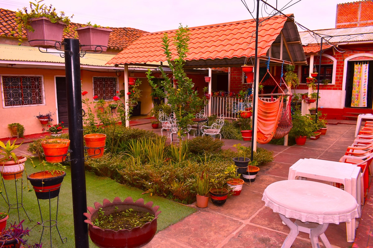 A charming garden area is surrounded by vibrant greenery and colorful potted plants. A covered patio features a tiled roof with white chairs and a hammock creating a relaxed atmosphere. Pathways intersect through the garden, providing easy access to different seating areas.