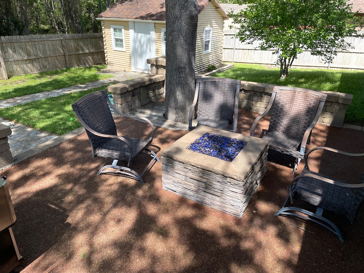 A cozy outdoor seating area features four comfortable chairs arranged around a fire pit. A stone wall encloses the fire pit, which is surrounded by finely crushed gravel. Tall trees provide shade, and a fenced backyard with a well-maintained lawn is visible in the background.