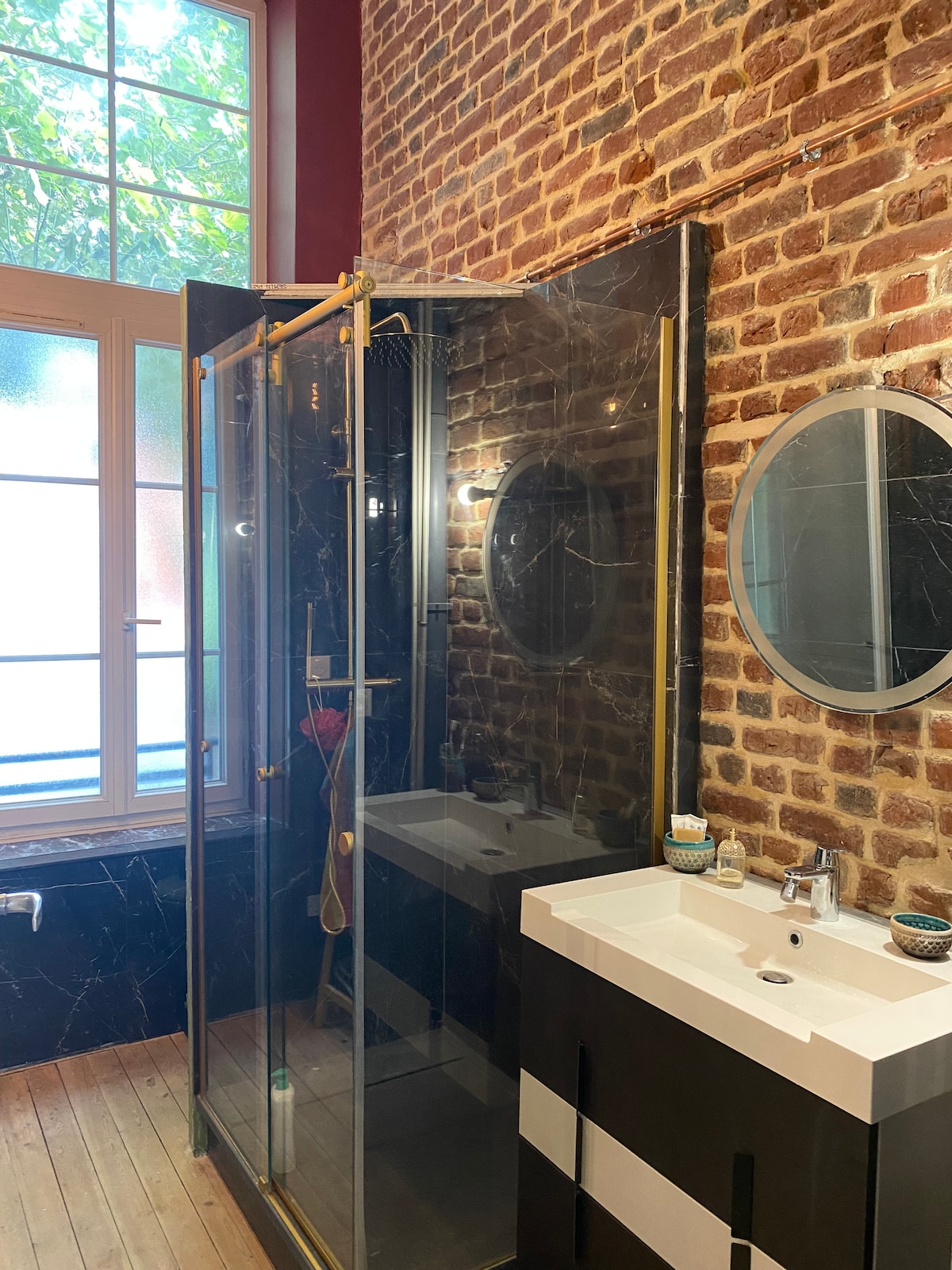 The bathroom features a walk-in glass shower enclosed by black framing. A modern vanity with a white countertop and a round mirror is positioned beside it. Exposed brick walls add texture, while natural light filters through a large window.