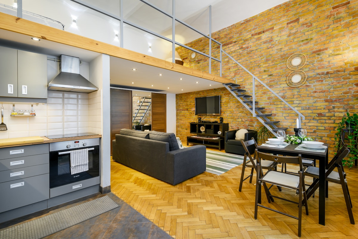 A modern, open-concept living area is featured with a cozy seating arrangement, complemented by a television. The kitchen, equipped with sleek cabinetry and an integrated oven, merges seamlessly with a dining space set for four. Exposed brick walls add texture to the stylish interior.