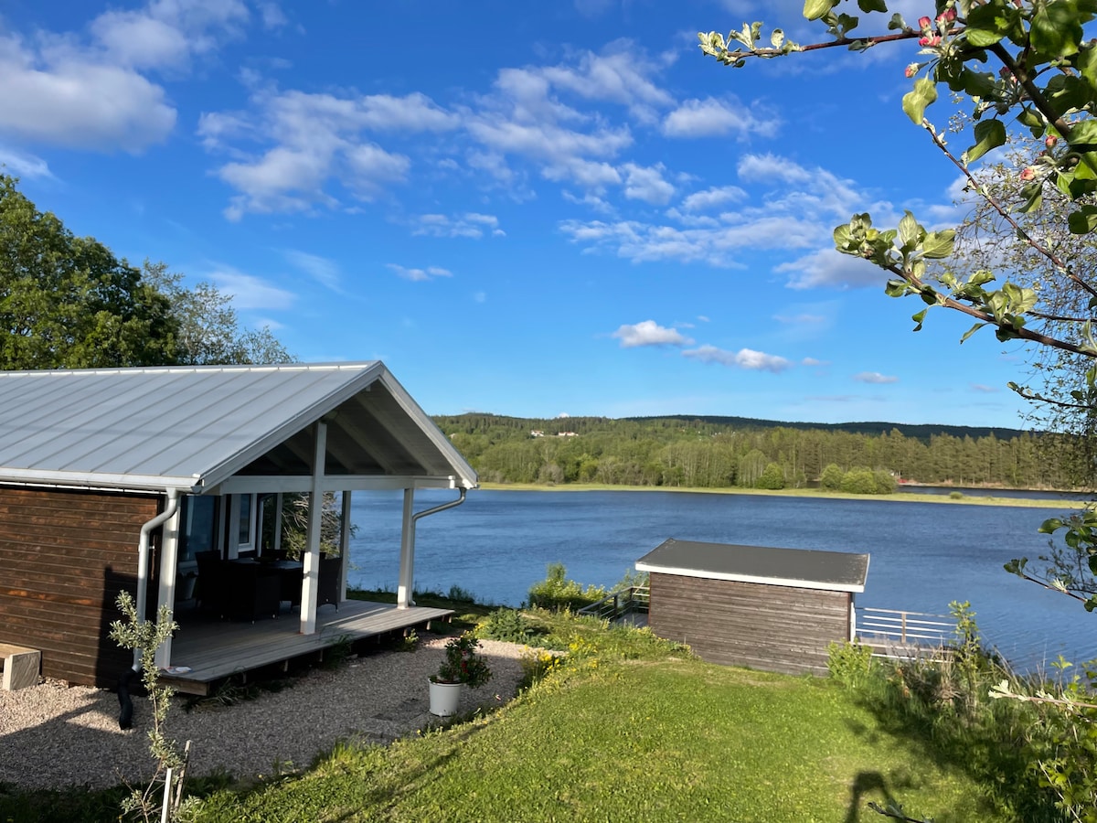 This image showcases a serene landscape featuring a wooden house with a covered porch overlooking the Ljungan river. Lush greenery and blooming flowers surround the area, while a small shed is positioned nearby. A clear blue sky complements the peaceful outdoor setting.