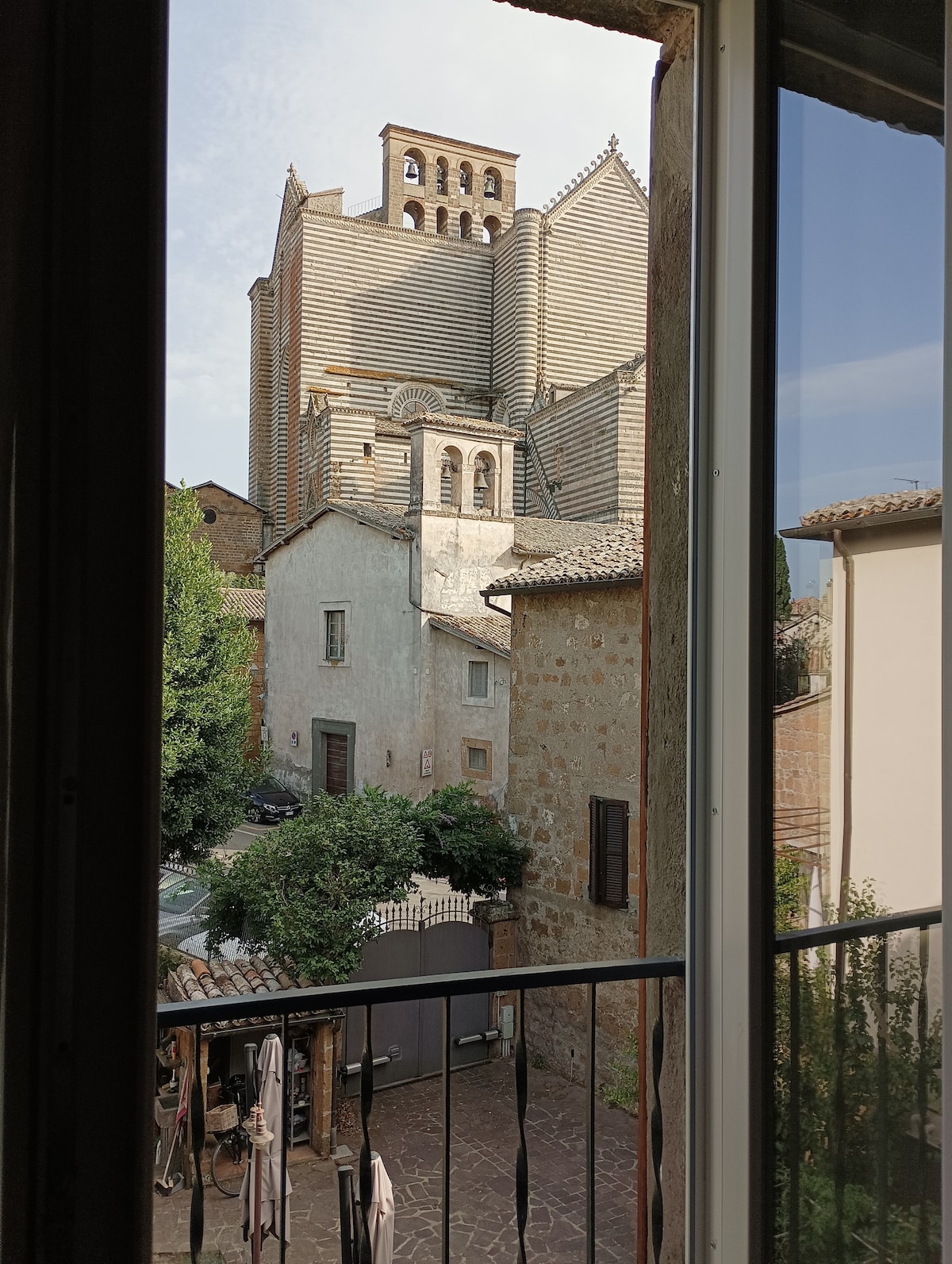 A view from the window reveals the exterior of a historic building, showcasing distinct architectural features. Nearby trees and quaint structures line the street below, while a small courtyard with outdoor seating is visible in the foreground.