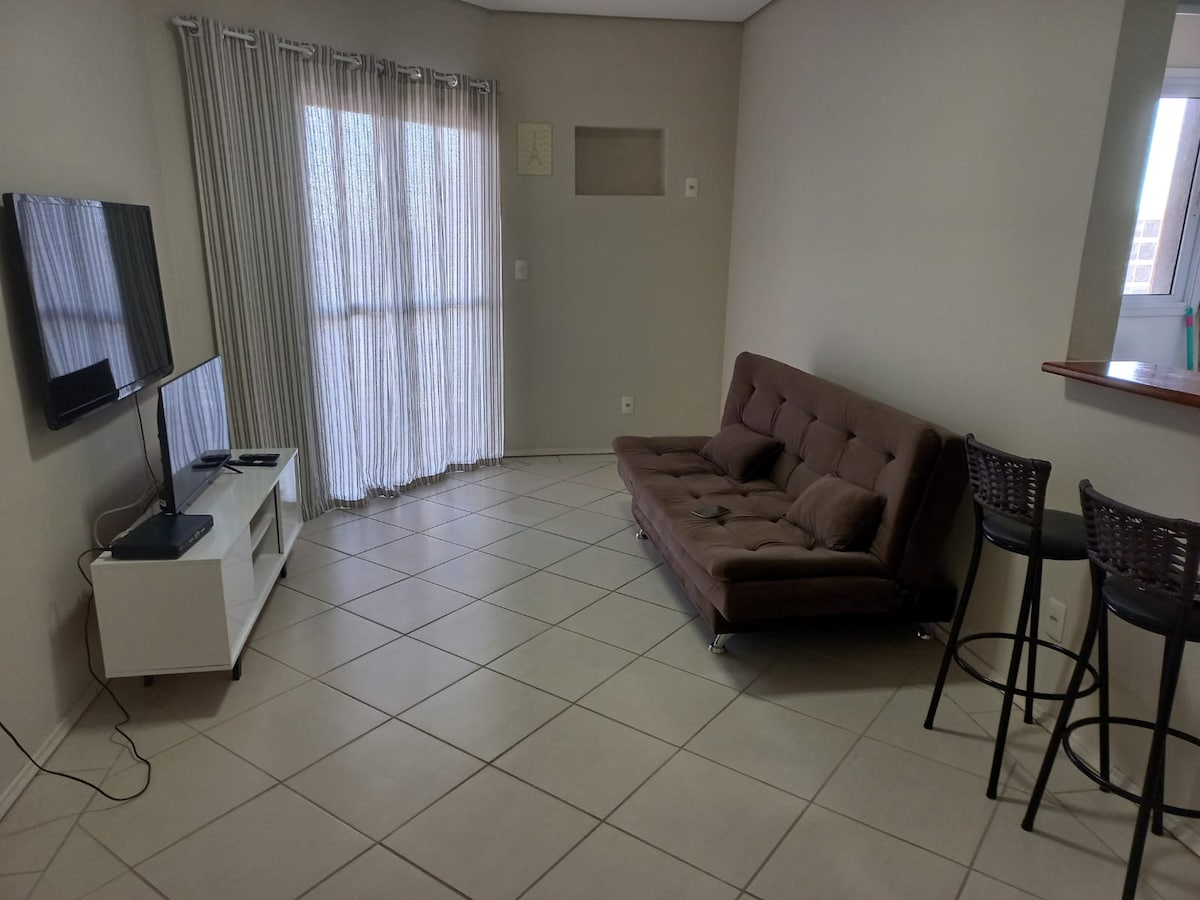 A spacious living area features a brown sofa bed facing a wall-mounted television. Natural light enters through sheer curtains covering the sliding glass doors. In the corner, two high bar stools are positioned by a counter, enhancing the functionality of the space.