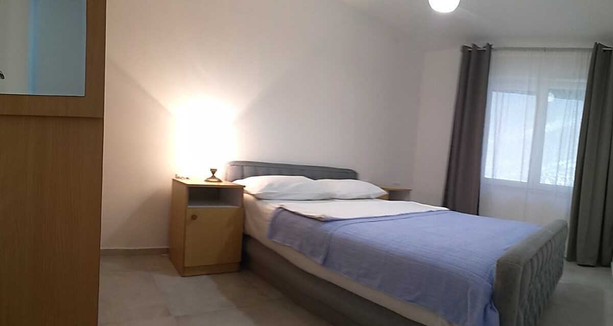 A spacious sleeping area features a comfortable bed with a light blue blanket neatly spread. A bedside table holds a soft lamp, providing gentle illumination. Natural light is welcomed through a window adorned with light curtains, complementing the neutral-toned walls.