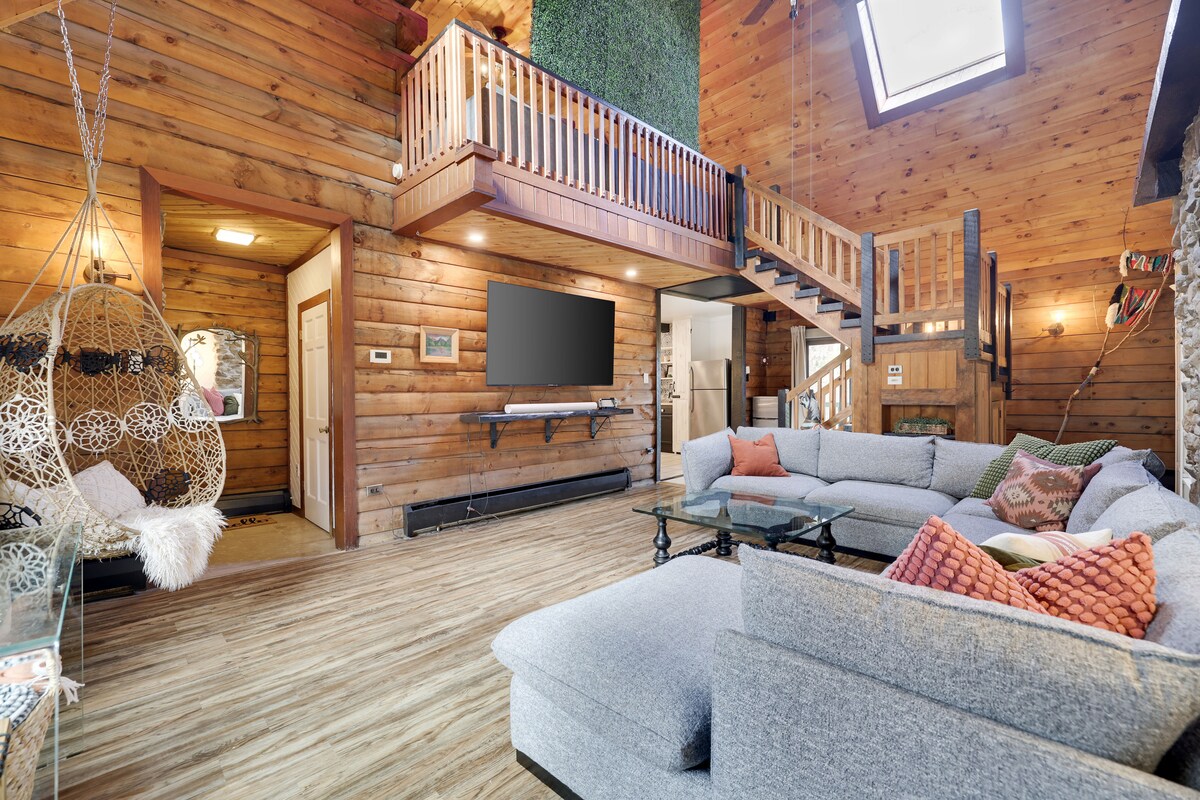 The interior living space features wood-paneled walls with exposed log beams and a spacious, cozy couch. A large television is mounted on the wall, while a unique hanging chair adds character. Skylights and large windows enable ample natural light, enhancing the open atmosphere.