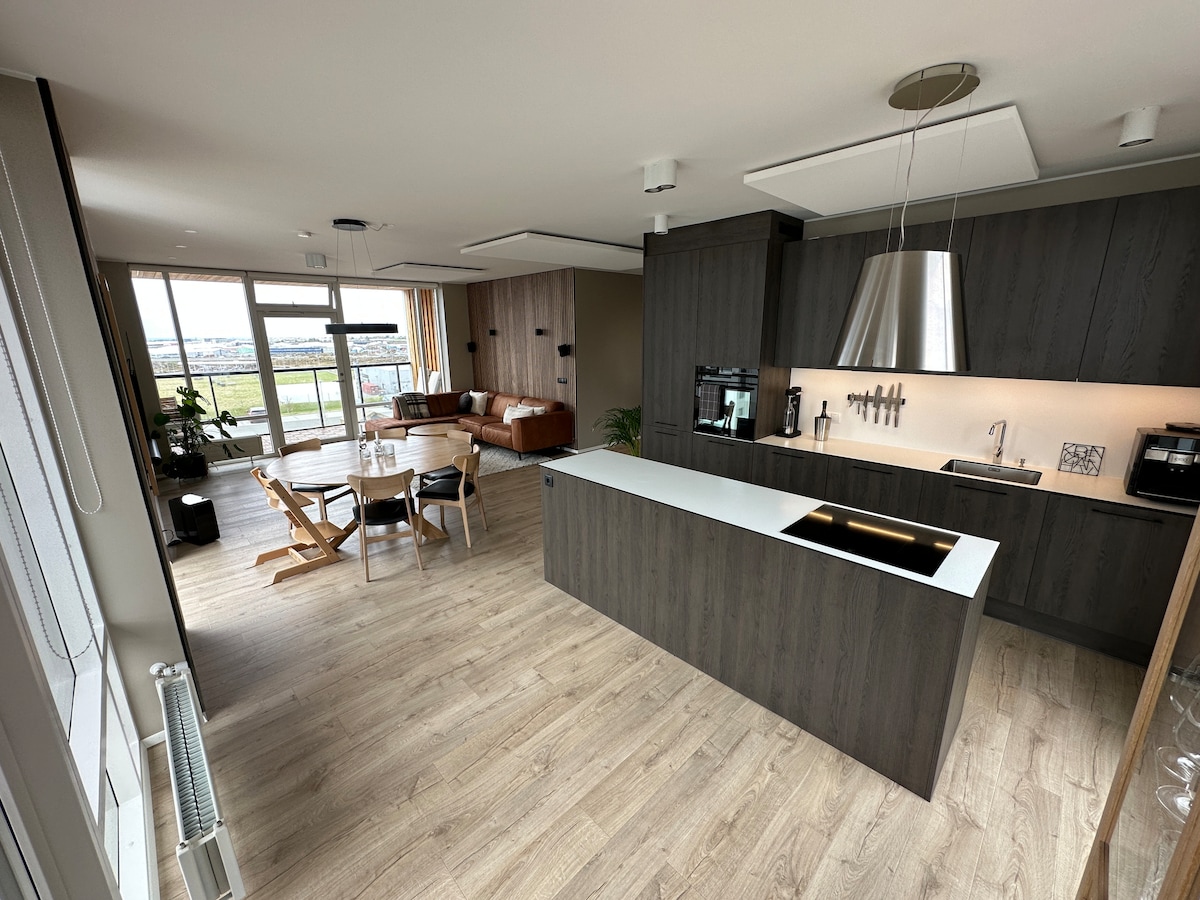 A modern kitchen and living area is displayed with sleek cabinetry and an island featuring a sink. Natural light enters through large windows. A dining table with four chairs is set nearby, and a cozy seating area with a sofa and coffee table is visible.