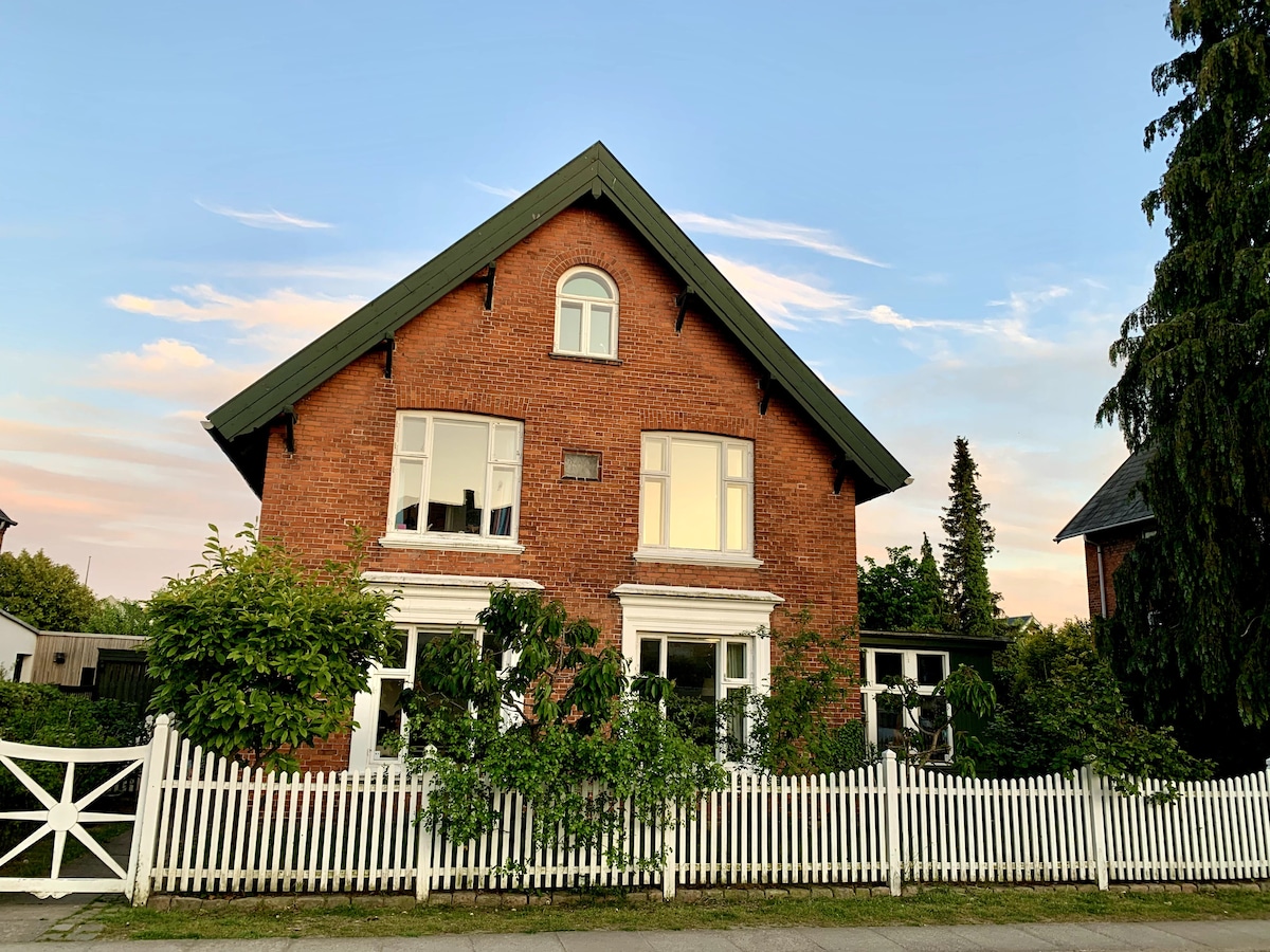A charming red-brick townhouse is set against a soft sky, featuring large windows framed by greenery. A white picket fence borders the property, enhancing its inviting exterior. The peaked roof adds character to the structure, with trees visible in the background.