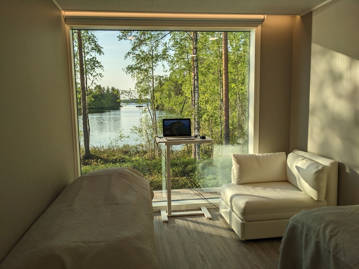 The room features large glass windows overlooking a serene lake framed by lush greenery. A workspace is provided with a modern desk, and a cozy seating area includes a sofa. The aesthetic is complemented by soft, neutral tones throughout the space.