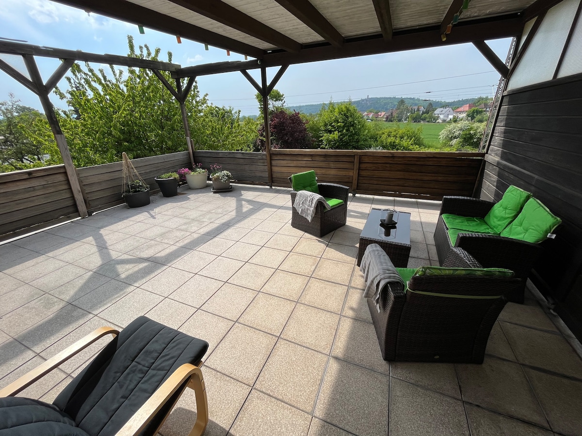 A spacious terrace features comfortable seating arrangements with green cushions and a low table, surrounded by potted plants. The area is shaded by a wooden roof, with views of greenery visible in the distance under a clear sky.