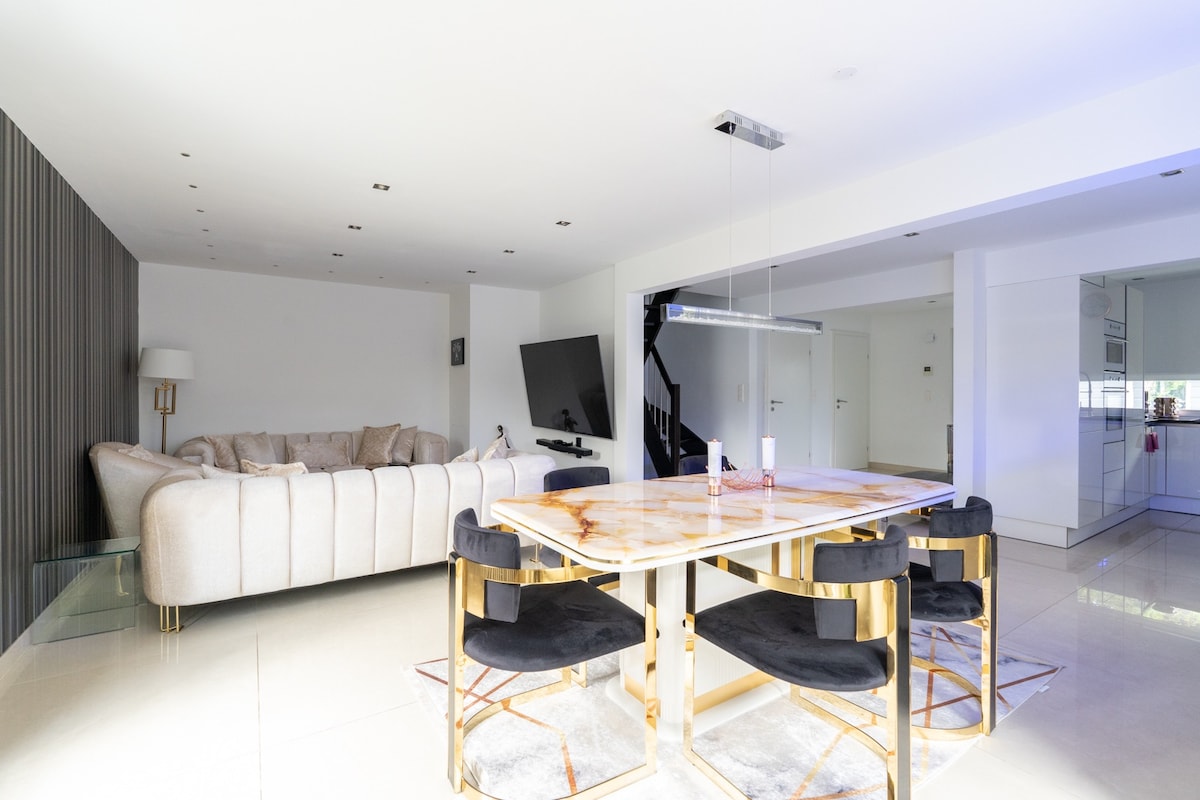 An airy living and dining area features a large cream-colored sectional sofa and a sleek dining table with a marble top. Black chairs with gold accents surround the table. Soft lighting enhances the modern aesthetic of the space.